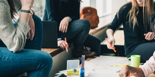 Vier Menschen sitzen an einem Tisch, die Gesichter sind nicht im Bild. Sie halten Stifte in der Hand und sehen nachdenkend aus. Eine Hand schreibt etwas auf das Plakat auf dem Tisch, welches sehr unstrukturiert gestaltet ist. Auf dem Tisch sind noch mehr Stifte und eine Tasse. Auf der Sitzbank erkennt man einen geöffneten Laptop und einen Rucksack im Hintergrund.