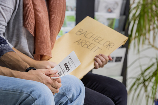 Zwei Personen sitzen nebeneinander. Man sieht einen Ausschnitt von Knie bis Brust. Die hintere Person hat einen großen Umschlag mit der Aufschrift „Bachelor-Arbeit“ in der Hand, die vordere Person hat einen kleinen weißen Zettel mit einer Wartenummer, einem Code und dem TU-Logo in der Hand.