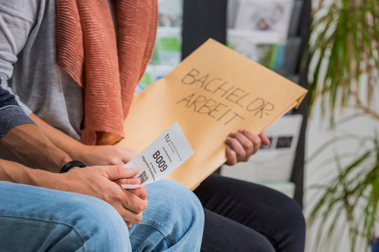 Zwei Personen sitzen nebeneinander. Man sieht einen Ausschnitt von Knie bis Brust. Die hintere Person hat einen großen Umschlag mit der Aufschrift „Bachelor-Arbeit“ in der Hand, die vordere Person hat einen kleinen weißen Zettel mit einer Wartenummer, einem Code und dem TU-Logo in der Hand.